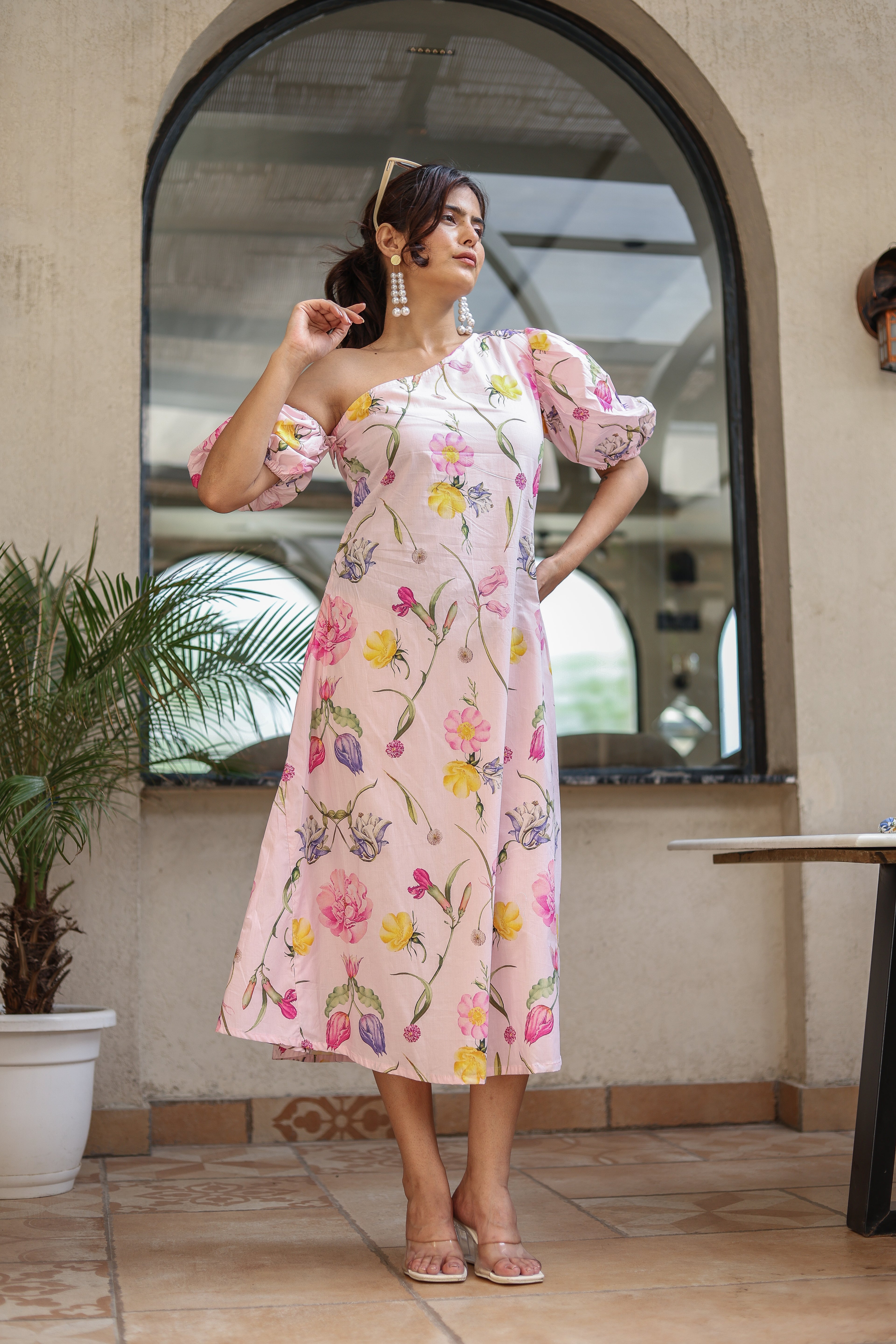 Woman in a floral dress standing in front of a mirror with a plant and table in the background.